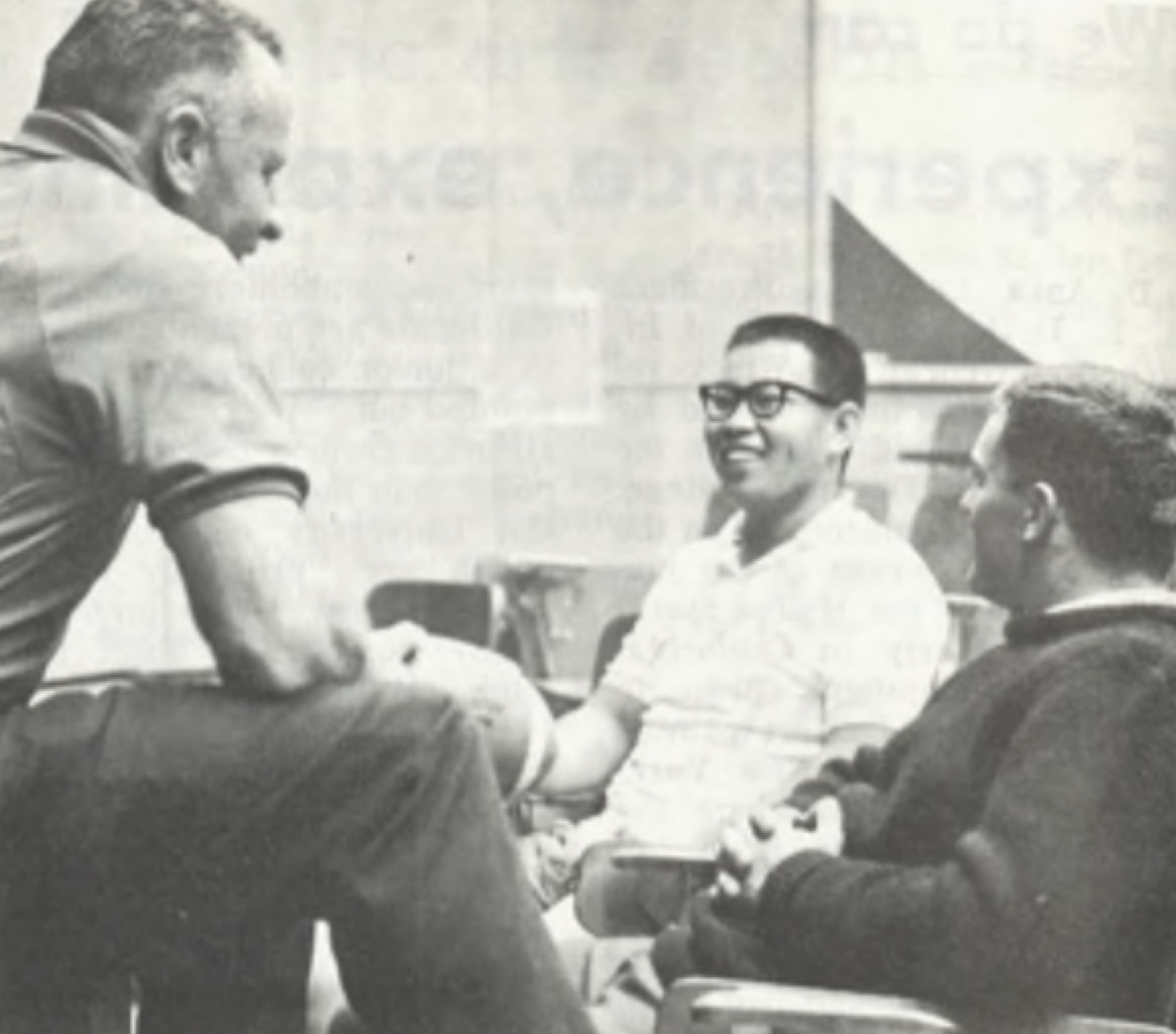 Two players talk to their coach during De Anza's inaugural football season (La Voz, 1967) Three White and Asian men are sitting and engaged in conversation. The man on the left is sitting on a table or elevated surface, while the other two are seated in chairs.