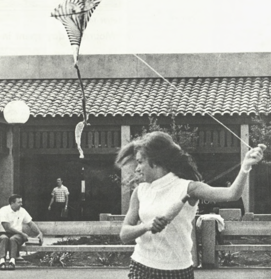 A De Anza student enjoys springtime (La Voz, 1969) A young woman in a sleeveless top and plaid skirt flies a kite outside a tiled-roof building, with a few people sitting on a bench in the background. Her hair is blowing in the wind as she enjoys a sunny day.