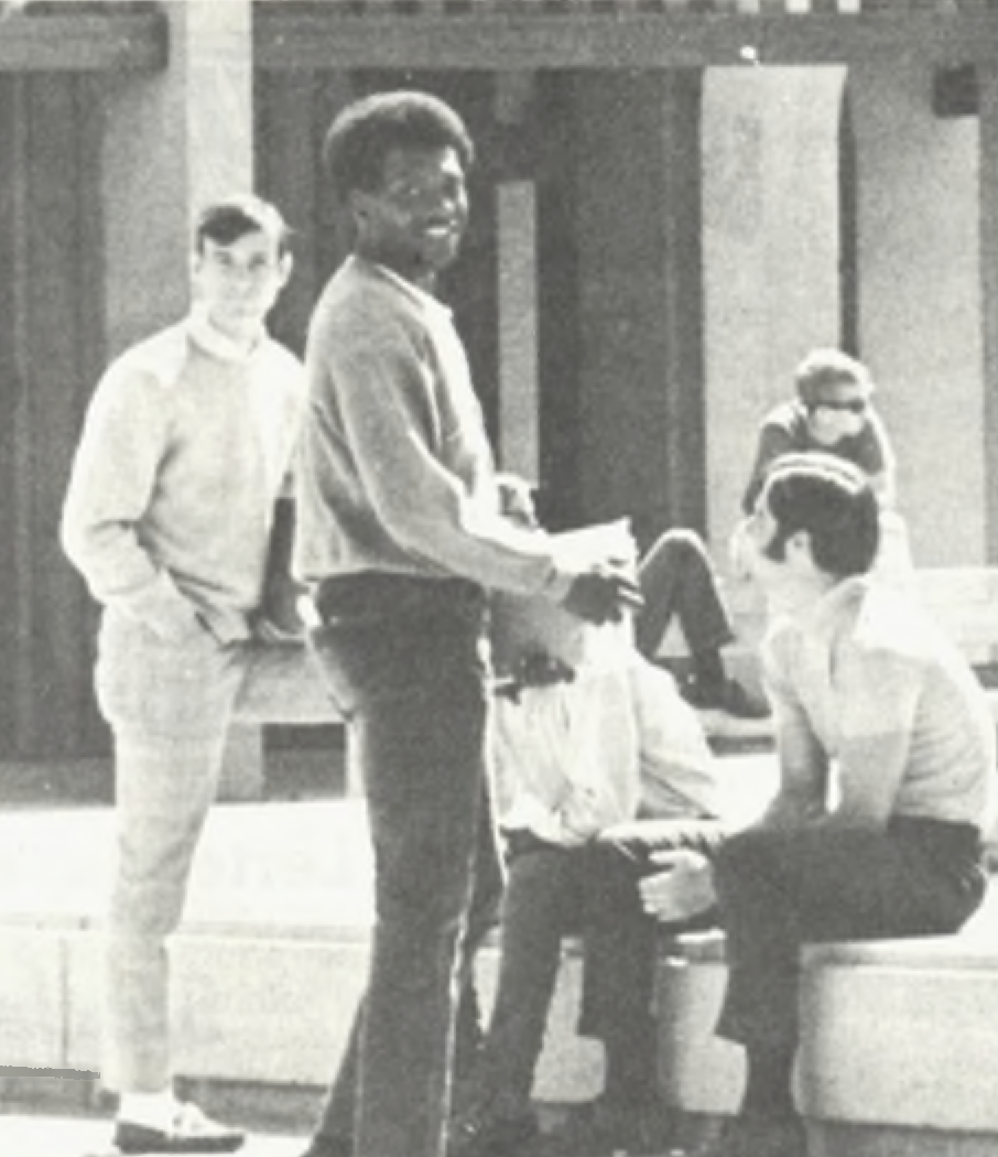Despite their relatively few numbers, Black and Chicano students formed strong student unions on campus (La Voz, 1968) A group of students is gathered in an outdoor setting, casually interacting. One of them, a young Black man, is standing holding papers and smiling at the camera, while others are seated on stone benches.
