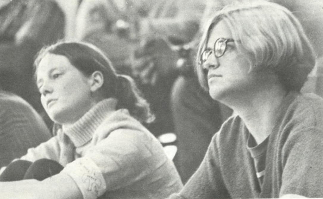 Two people seated and attentively listening. One has long dark hair and a turtleneck, while the other has short light hair and glasses. 