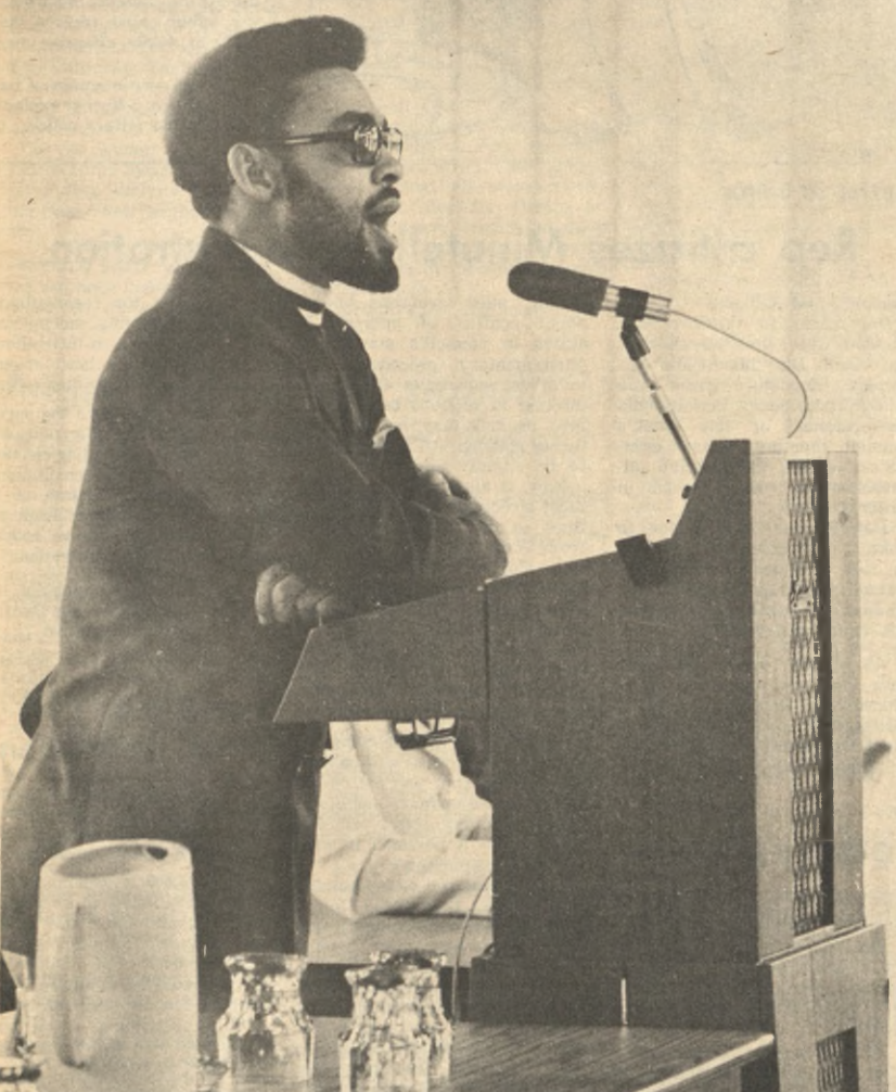 A Black man with glasses and a beard speaks passionately at a podium with a microphone.