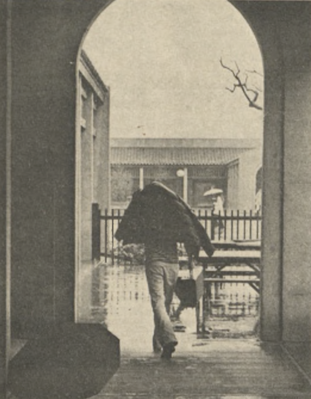 A person walks through an archway on a rainy day, holding a coat over their head. The ground is wet, and another figure holding an umbrella is visible in the background, near a gate and a leafless tree.