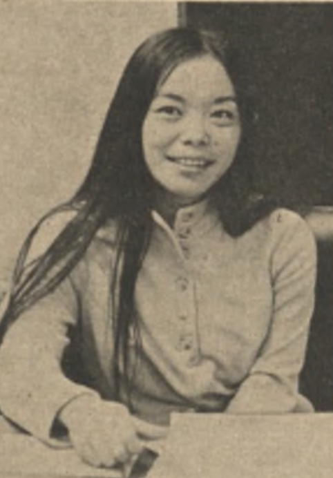A black and white photo of an Asian woman with long dark hair, seated and smiling at the camera. She is wearing a long-sleeved button-up shirt. 