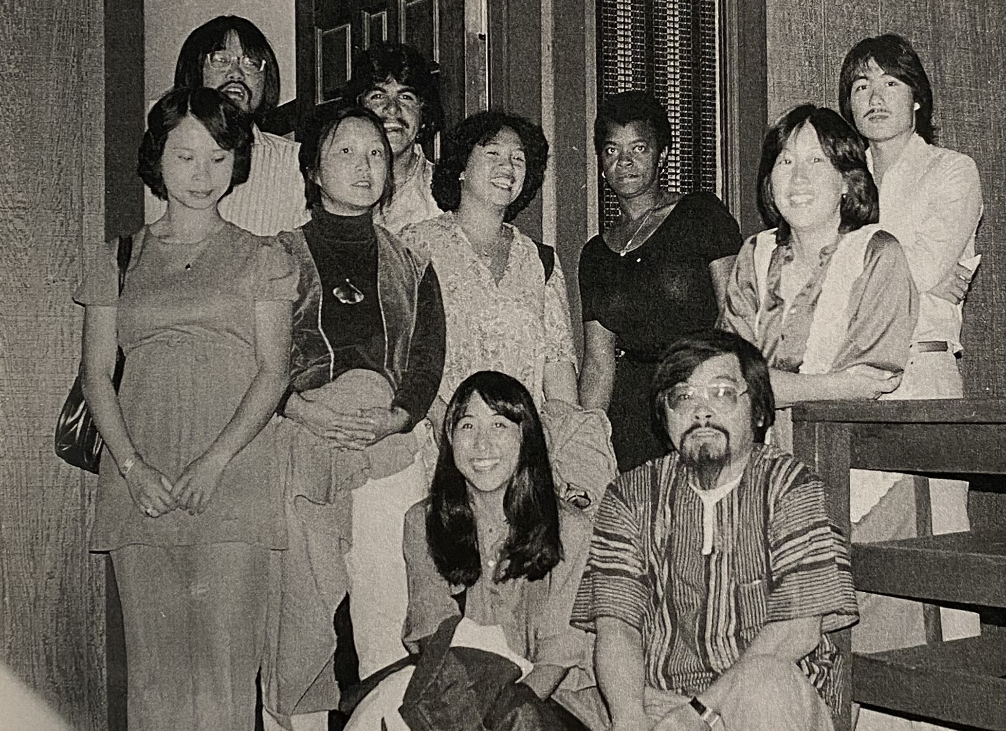 A group of diverse people posing together indoors, some sitting and some standing, with a staircase visible. They are wearing a variety of casual attire and appear to be in a friendly, relaxed setting.