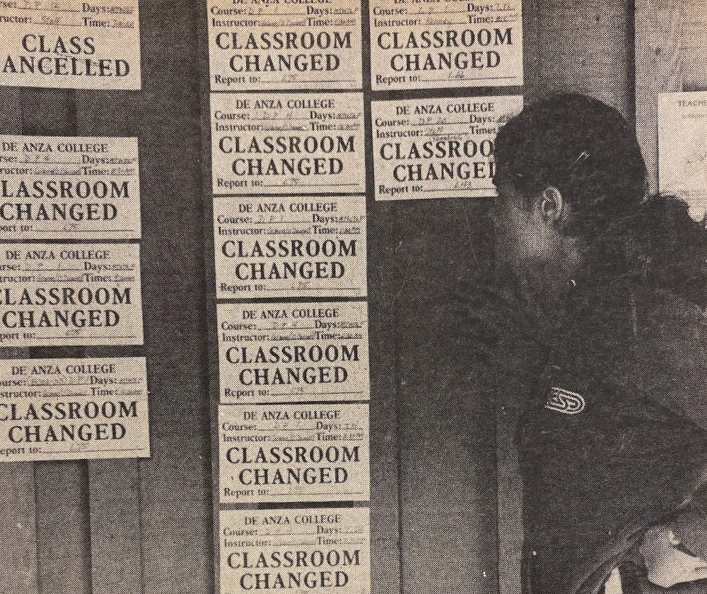 A person looks at a bulletin board filled with notices about classroom changes and class cancellations at De Anza College. The notices are printed on paper and cover most of the board.