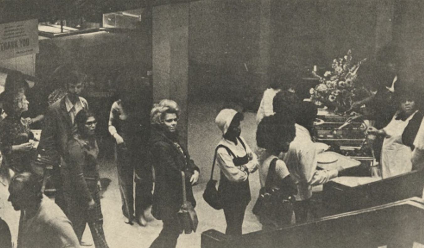 Show thumbnail preview A black and white photo showing a cafeteria scene. People are lined up to get food, served by staff behind a counter. Some individuals wear coats, while others carry trays. 