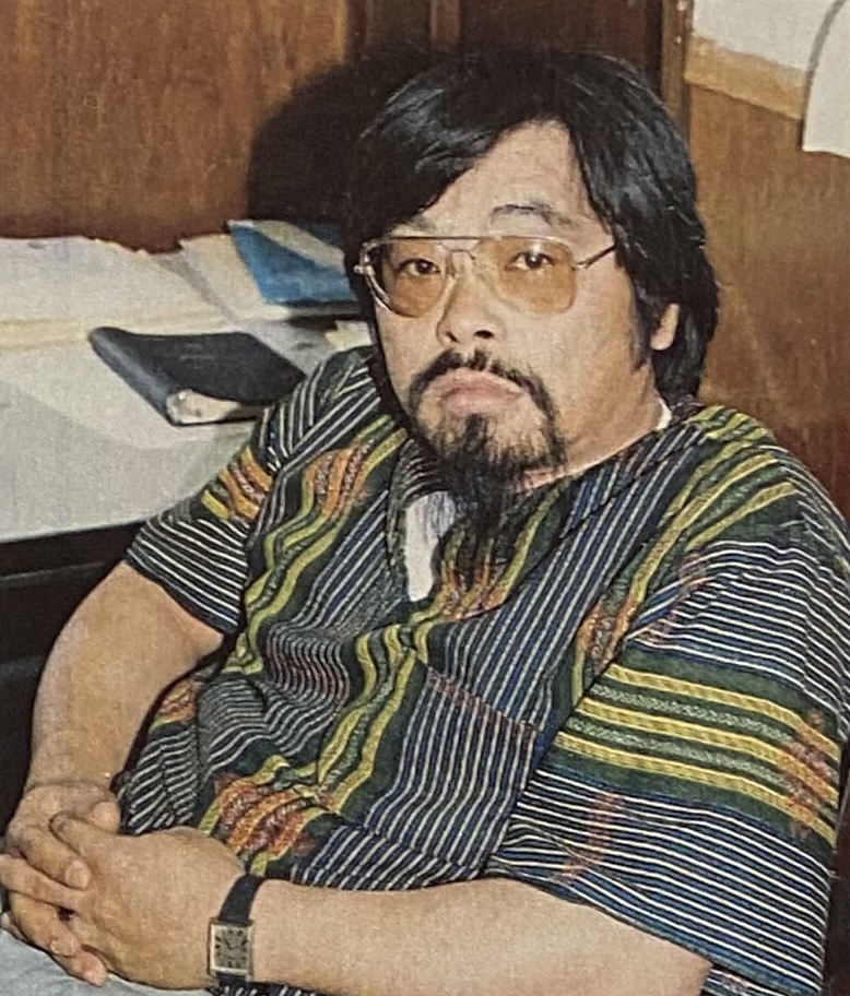 An Asian man with glasses and a beard sits in a wooden-paneled room, wearing a colorful striped shirt. He is seated with hands clasped on his lap, looking toward the camera. 
