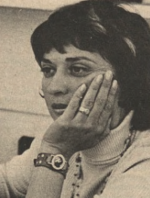 A woman with short dark hair, wearing a turtleneck sweater and a wristwatch, rests her chin on her hand, looking thoughtfully to the side.