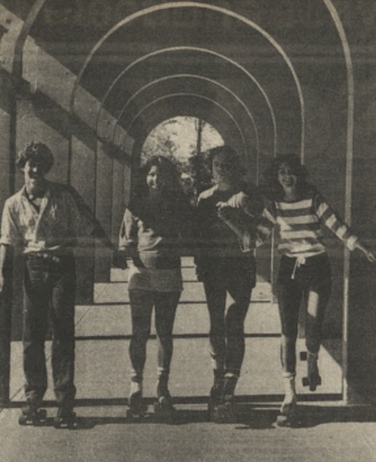 Four people roller skate under an arched pathway. They are side by side, smiling and enjoying the sunny day. 