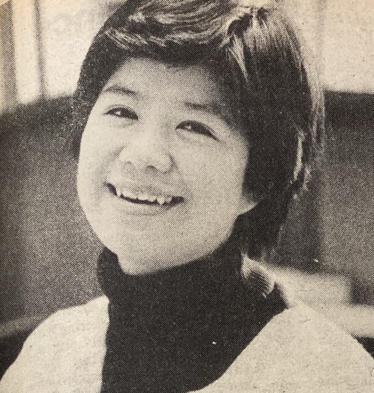 Black and white image of a young Asian woman with short hair, smiling. They are wearing a dark turtleneck under a light top. 