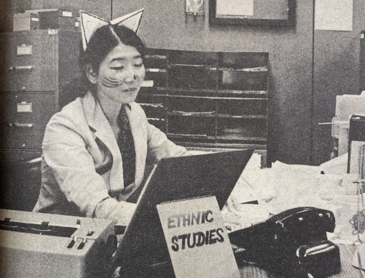 An Asian woman wearing cat ears and face paint resembling a cat works at a desk in an office. She is using a typewriter and have papers and files around. A sign reading Ethnic Studies is visible on the desk.