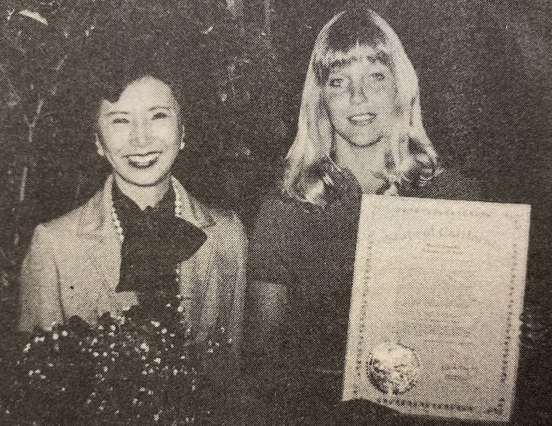 Two women smiling, one holding a bouquet of flowers and the other holding a document. Both are dressed in formal attire. 