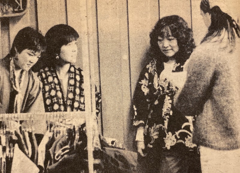 Designers display their creations during an Asian Awareness Month fashion show. (La Voz, 1982) Four Asian women are gathered indoors, engaging in conversation and standing in front of a clothing rack