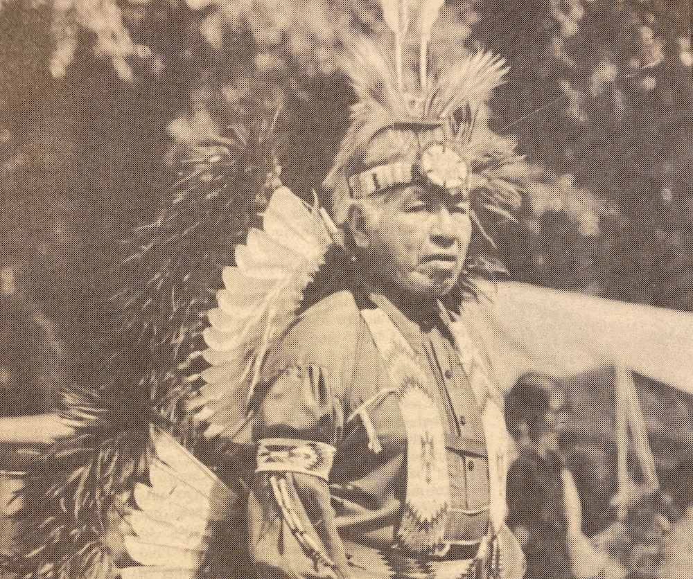 A Native American man wearing traditional regalia stands outdoors, adorned with intricate feather headdresses and armbands