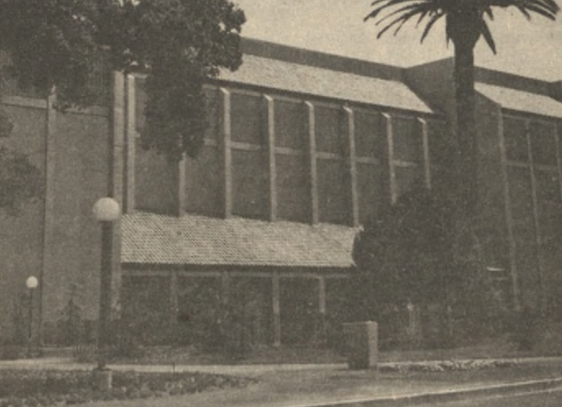 Vintage photograph of a building with large windows and a tiled roof, surrounded by trees and a street lamp in front.