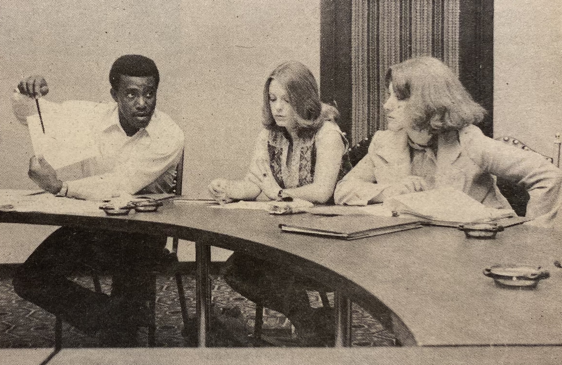 A Black man and two White women sit at a curved conference table in discussion. The man holds up a sheet of paper, gesturing while talking. The women are listening attentively, with one taking notes. Papers and folders are spread out on the table.