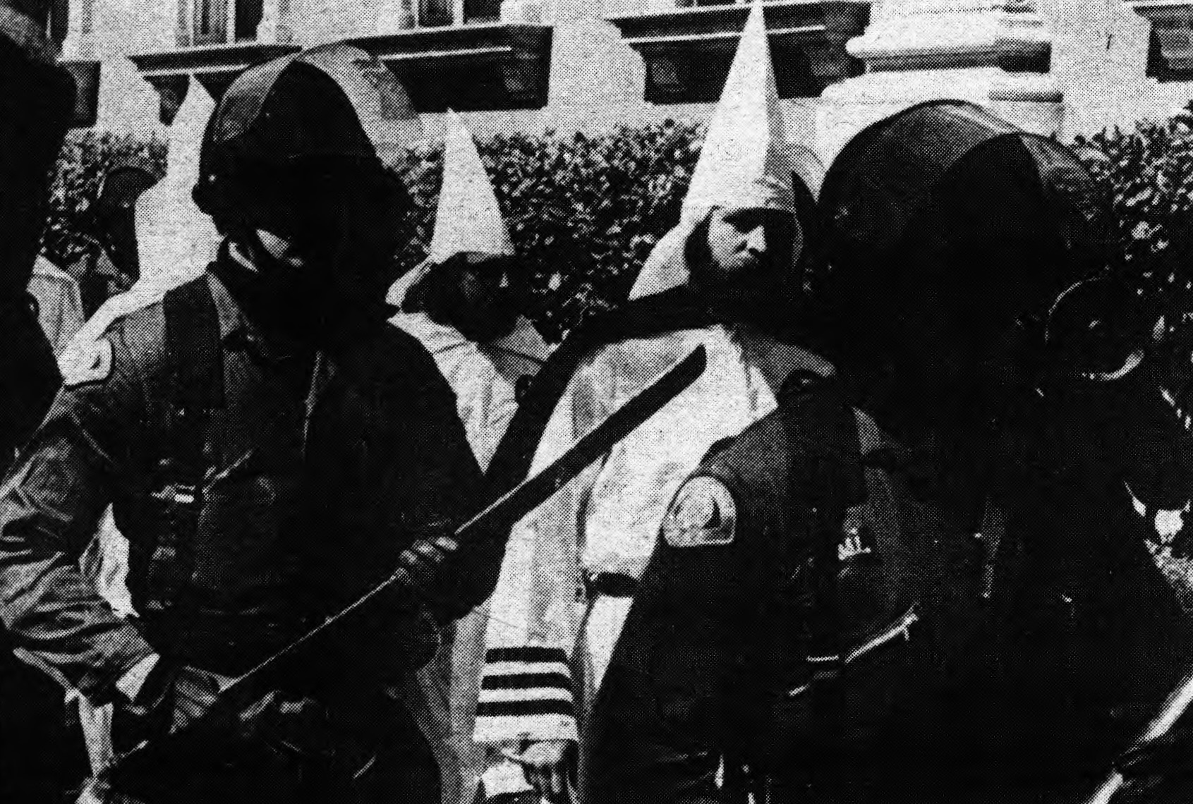 A black-and-white photo showing police officers in riot gear, including helmets and batons, standing in front of people wearing white robes and hoods. 