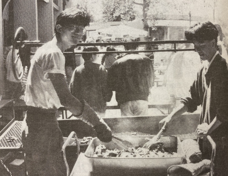 Members of the Korean Student Union hold a Korean Food Sale on the Campus Center patio as part of Asian Pacific Heritage Month. (La Voz, April 1991) Two Asian students are preparing food in large metal trays at an outdoor event. One person is stirring with a large spoon, while the other appears to be mixing ingredients.