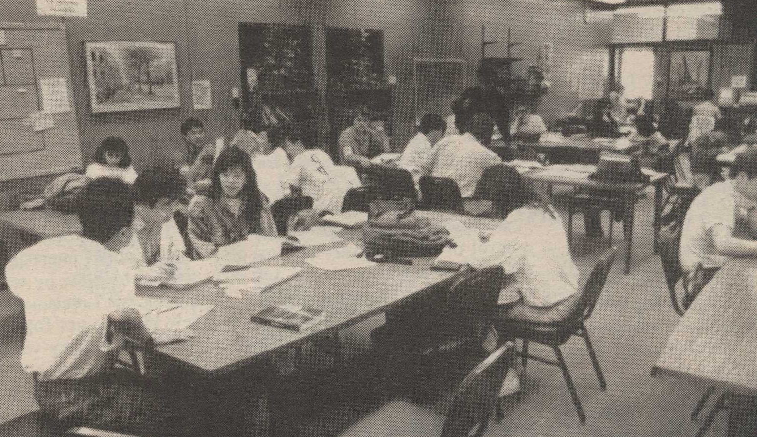 Tutorial center (La Voz, 1990) A group of students study in a classroom with rectangular tables and chairs. Some are reading, others are writing and discussing among themselves.