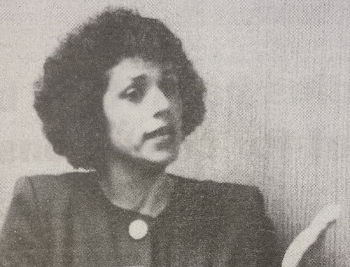 Lydia Ledesma speaks out in favor of IIS at a board meeting. (La Voz, 1993) Black and white image of a woman with curly hair, wearing a dark jacket with a round pin on the collar, who is currently engaged in a speech