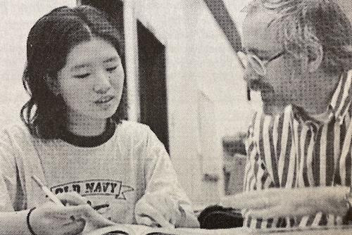 ESL (English as a Second Language) student and tutor. (La Voz, 1998) A black and white image of a woman and an older man sitting at a table. The woman is writing and appears to be explaining something to the man, who is wearing glasses and a striped shirt. Both are looking at papers on the table.