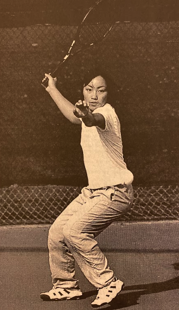 Tam Nguyen, a key player on the undefeated Women's Tennis State Championship team, trains in preparation for a repeat run at the state title. (La Voz, 1999) An Asian student in a white t-shirt and light pants is playing tennis on a court. Their stance is focused, holding the racket with both hands, ready to strike.