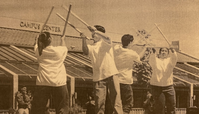 Members of PUSO (Philipino United Student Organization) perform a traditional Philipino dance. (La Voz, 1997) Four people perform a dance with wooden sticks in front of a building labeled Campus Center.