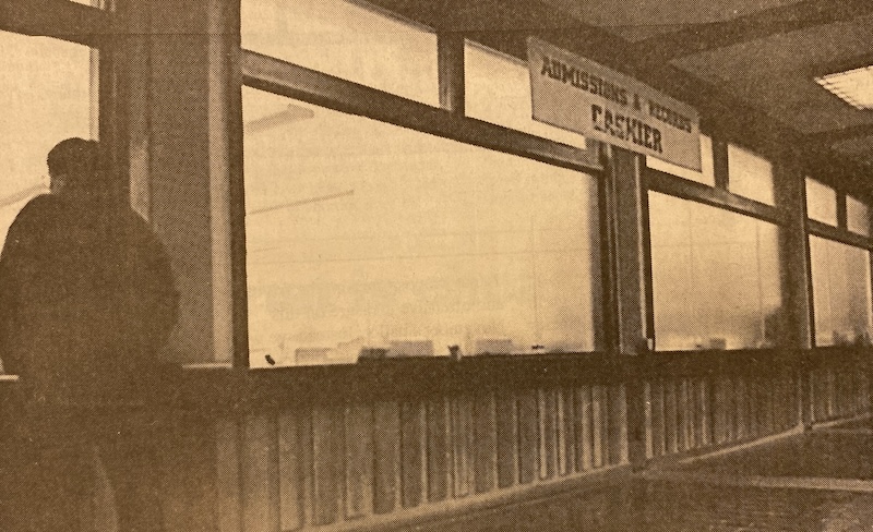 Cashier's office. (La Voz, circa 1990) A person stands inside a building, looking out through large windows. A sign reading Admissions & Business Cashier hangs above.
