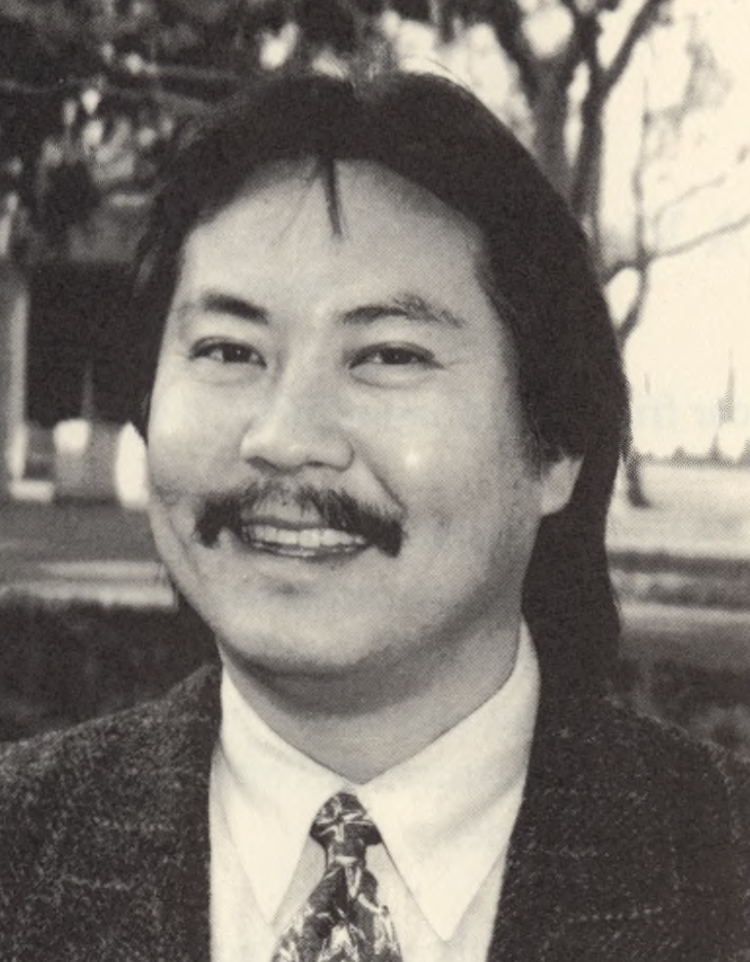 Duane Kubo (APALI opening ceremony program, 1997) A black and white photo of a person with long hair and a mustache, wearing a suit jacket, white shirt, and patterned tie.