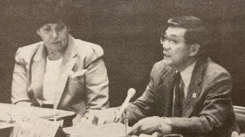 Dr. Betty Pacheco and Norman Mineta take part in the conference. (La Voz, 1991) Two people in formal attire are seated at a table with microphones, engaged in discussion. The person on the left wears a suit and the person on the right is speaking while gesturing with one hand.