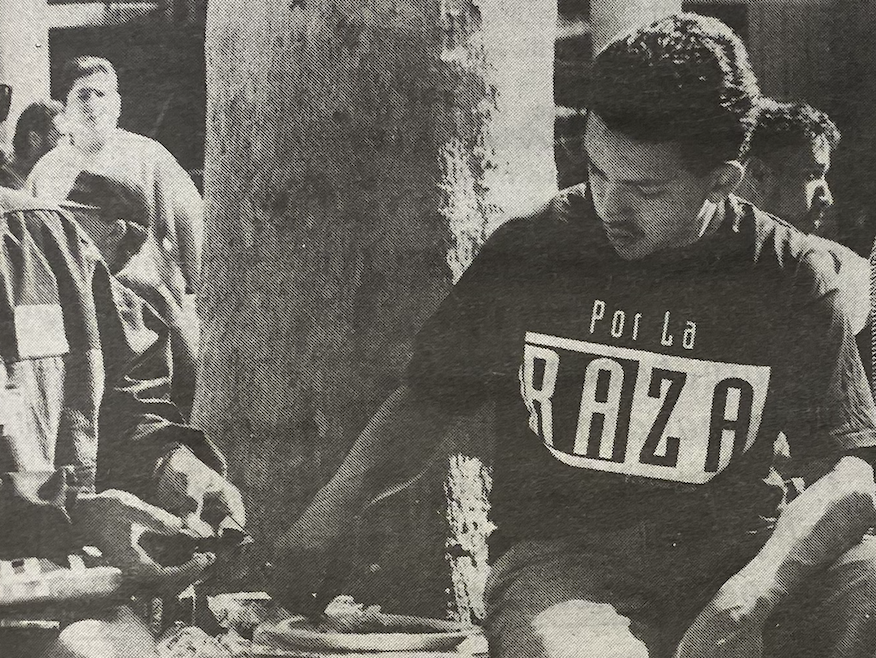 Students gather in front of the Hinson Campus Center for the La Raza/Latino Conference. (La Voz, February 1992) A black and white photo shows a young Latino man sitting near a tree, wearing a T-shirt with Por La Raza printed on it.