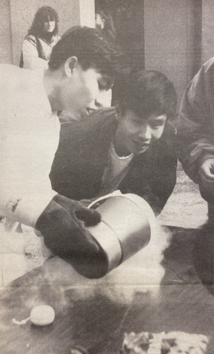 Students participate in Club Day activities. (La Voz, 1993) Two Asian students are participating in a science activity. One person is wearing gloves and pouring liquid from a container, creating vapor. The other person watches with interest. A small crowd observes in the background.