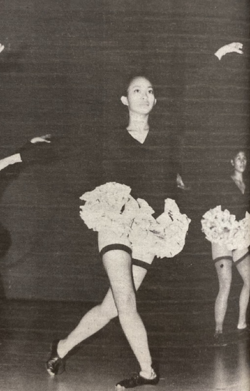 A black and white photo of a young Asian dancer on stage mid-performance. The dancer is wearing a short, ruffled skirt and is striking a graceful pose with one arm raised and one leg extended.