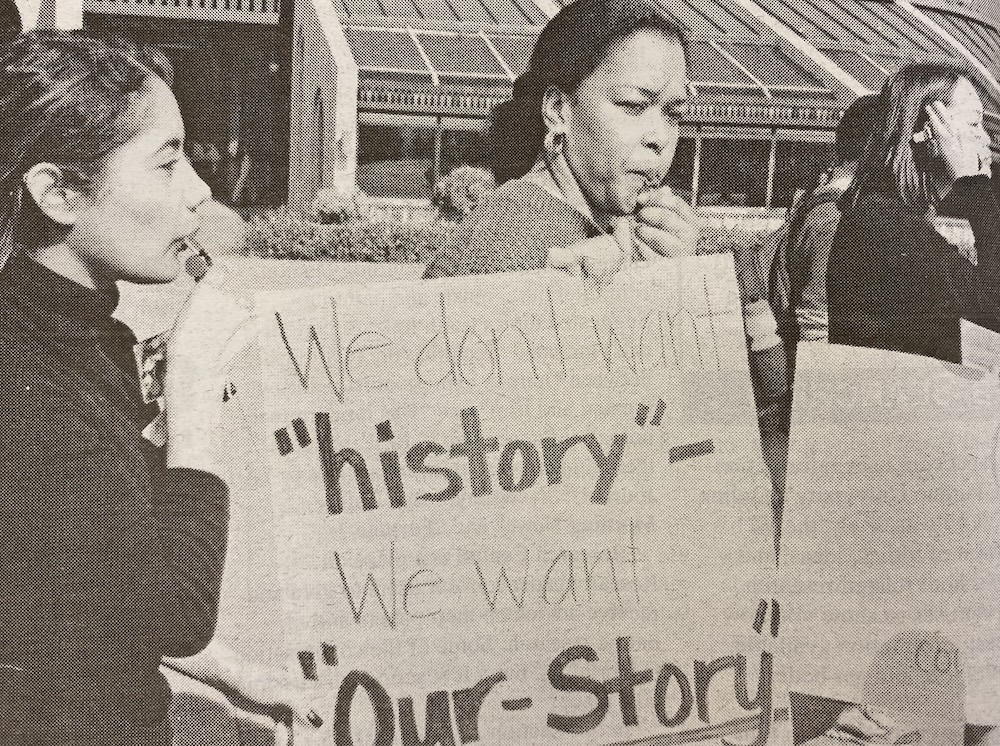 Two people hold a protest sign that reads: We don't want "history" - We want "Our-Story". They are standing outside, and one person is speaking into a megaphone.