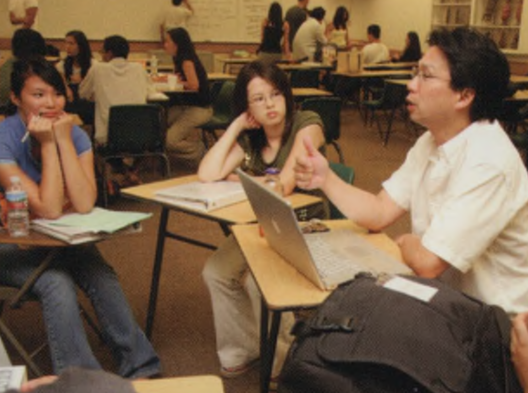 A group of students in a classroom sit at desks engaged in conversation. One student gestures with his hand while holding a laptop. 