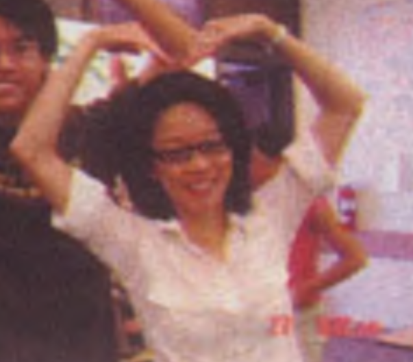A person with glasses, wearing a white shirt, makes a heart shape with their arms above their head.