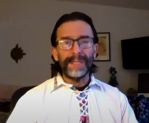 A man with glasses is wearing a white shirt with a decorative collar. He is sitting indoors, with a wall decoration and a framed picture visible in the background.