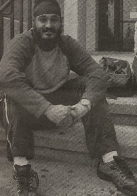 A South Asian student wearing a turban and casual clothing sits on stone steps, smiling with hands clasped. A backpack rests nearby.