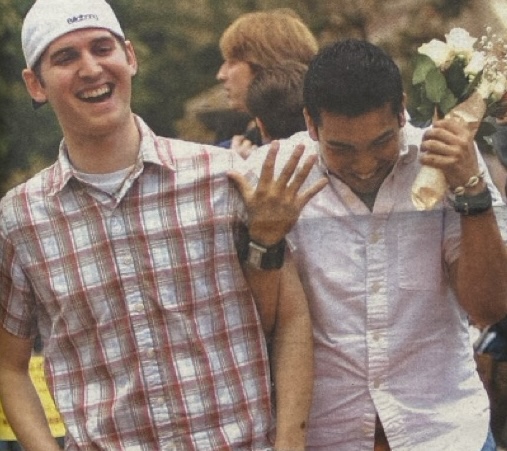 Two people walking and laughing together. One is wearing a white cap and plaid shirt, and the other is holding a bouquet.