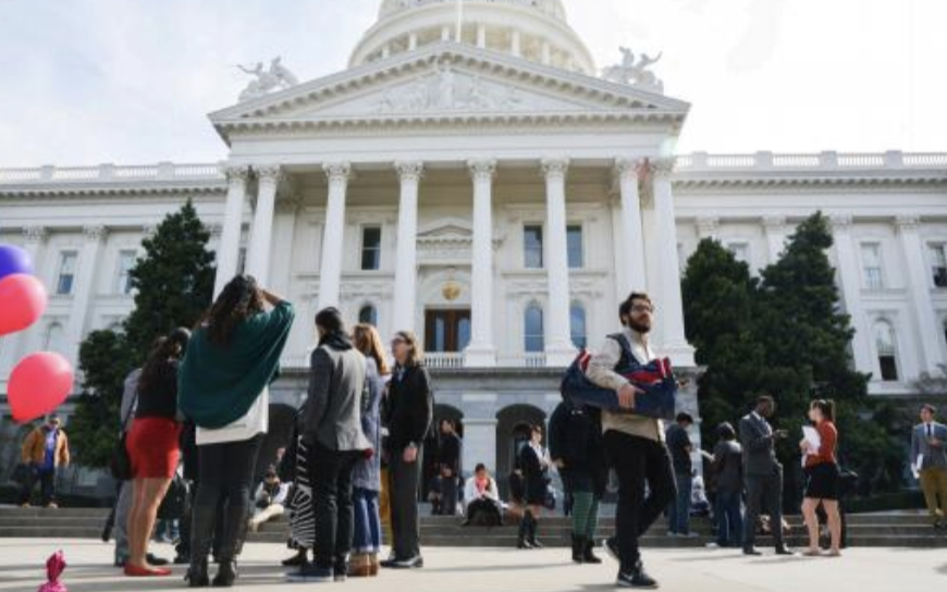 A group of people gather in front of a large government building with white columns and a dome. 