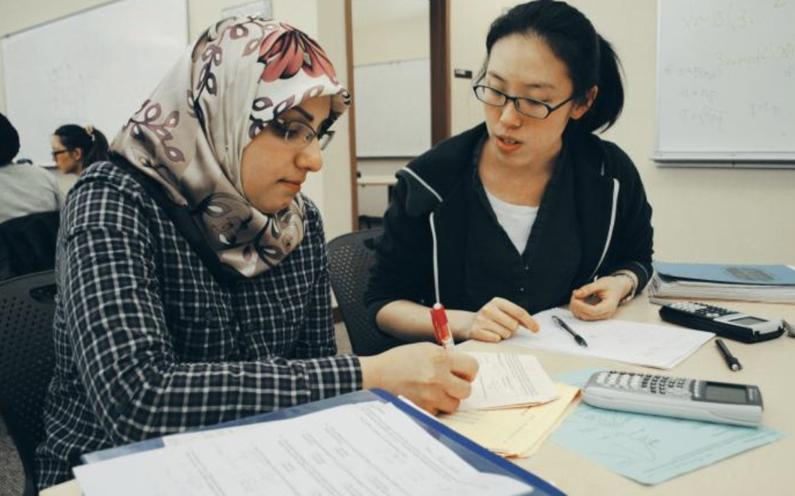 Two Arab and Asian American students are seated at a table, discussing papers and notes spread out in front of them. 