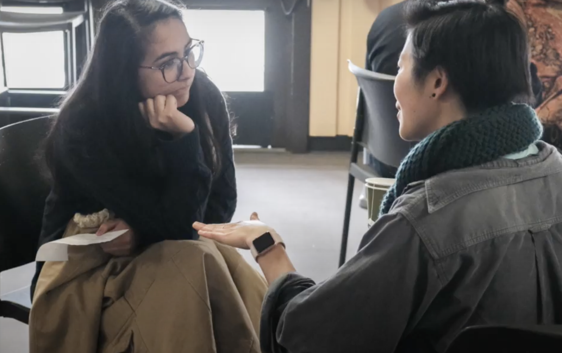 Two people are sitting in chairs, engaged in conversation. The person on the left has glasses and is resting their chin on their hand, while holding a piece of paper. The person on the right is gesturing with their hand, wearing a scarf and a watch.
