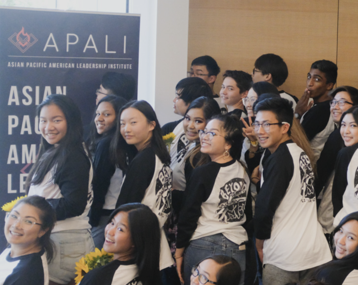 Show thumbnail preview A group of young people poses together for a photo, many wearing matching black and white shirts. They stand beside an APALI sign, which stands for Asian Pacific American Leadership Institute. Some hold sunflowers, smiling and looking towards the camera.