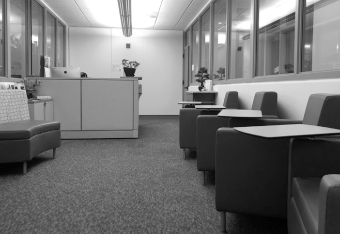 A black and white photo of a waiting room with a row of cushioned chairs and small attached tables on the right side. A reception desk with plants in the background is on the left. The room is lit by overhead lights.