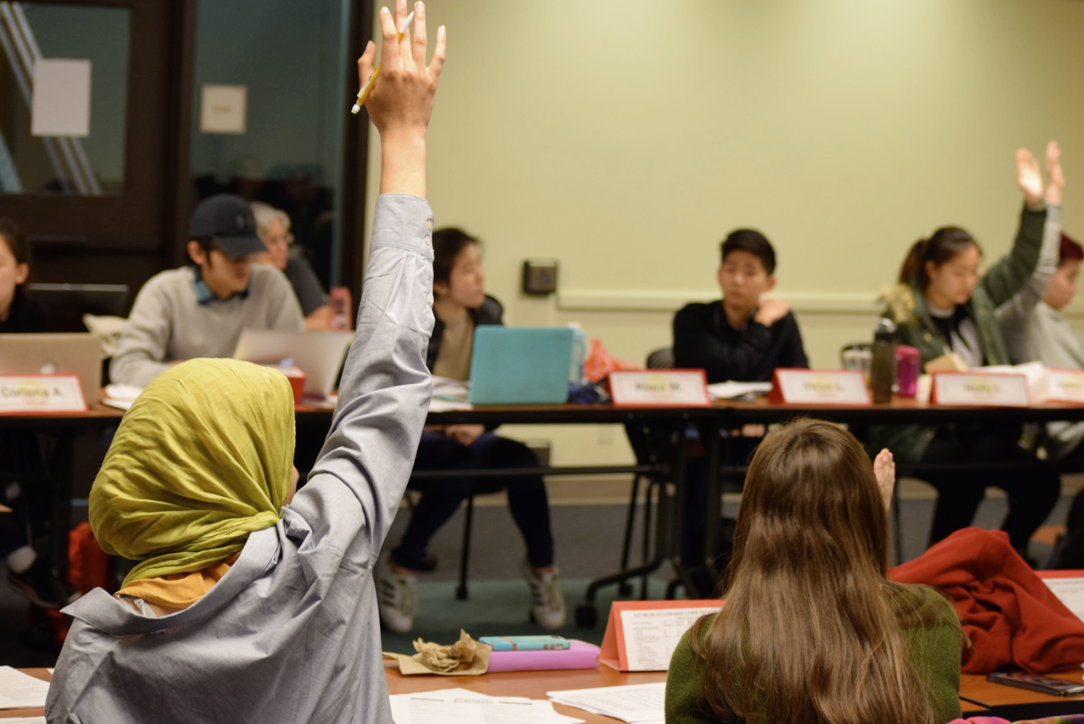 A classroom scene with students sitting around tables, some raising their hands. The focus is on a student wearing a green headscarf raising their hand, while others are using laptops and taking notes. 