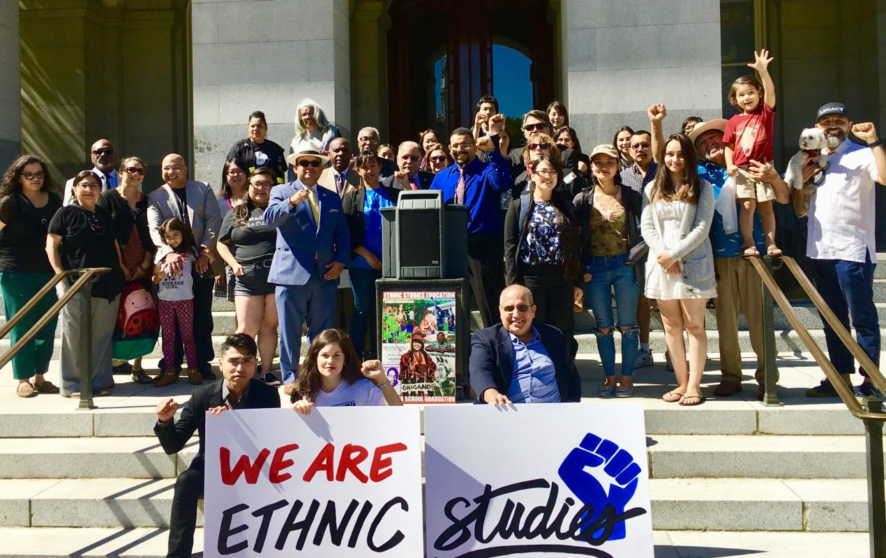A diverse group of people stand on steps holding signs reading We Are Ethnic Studies with a raised fist symbol. Several children are present, and a speaker stand is in front of the group.
