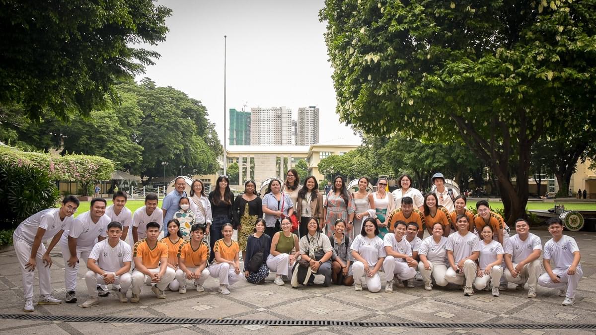 A large group of people posing outdoors in front of tall buildings and trees. Some are wearing white uniforms, others orange shirts, and others casual clothing. They are smiling and arranged in multiple rows on a plaza 