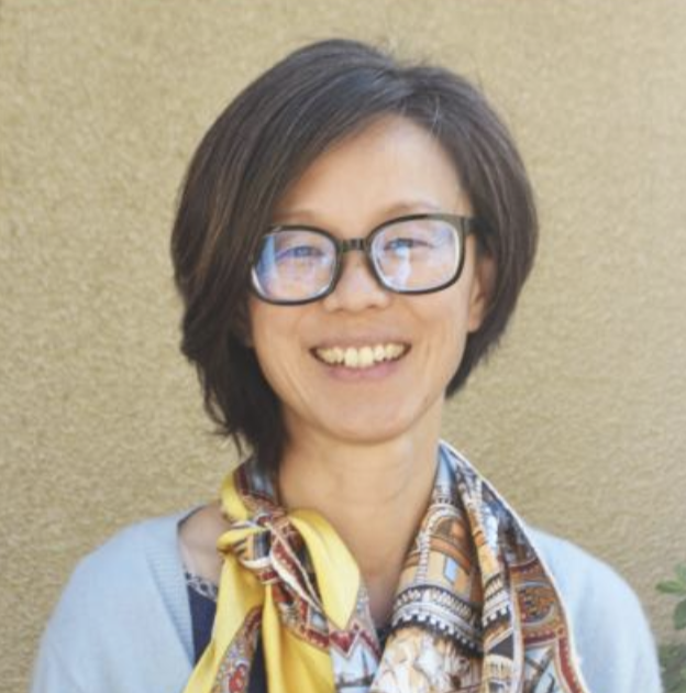 A person with short brown hair and glasses smiles at the camera. They are wearing a colorful scarf and a light blue top, standing against a beige wall.