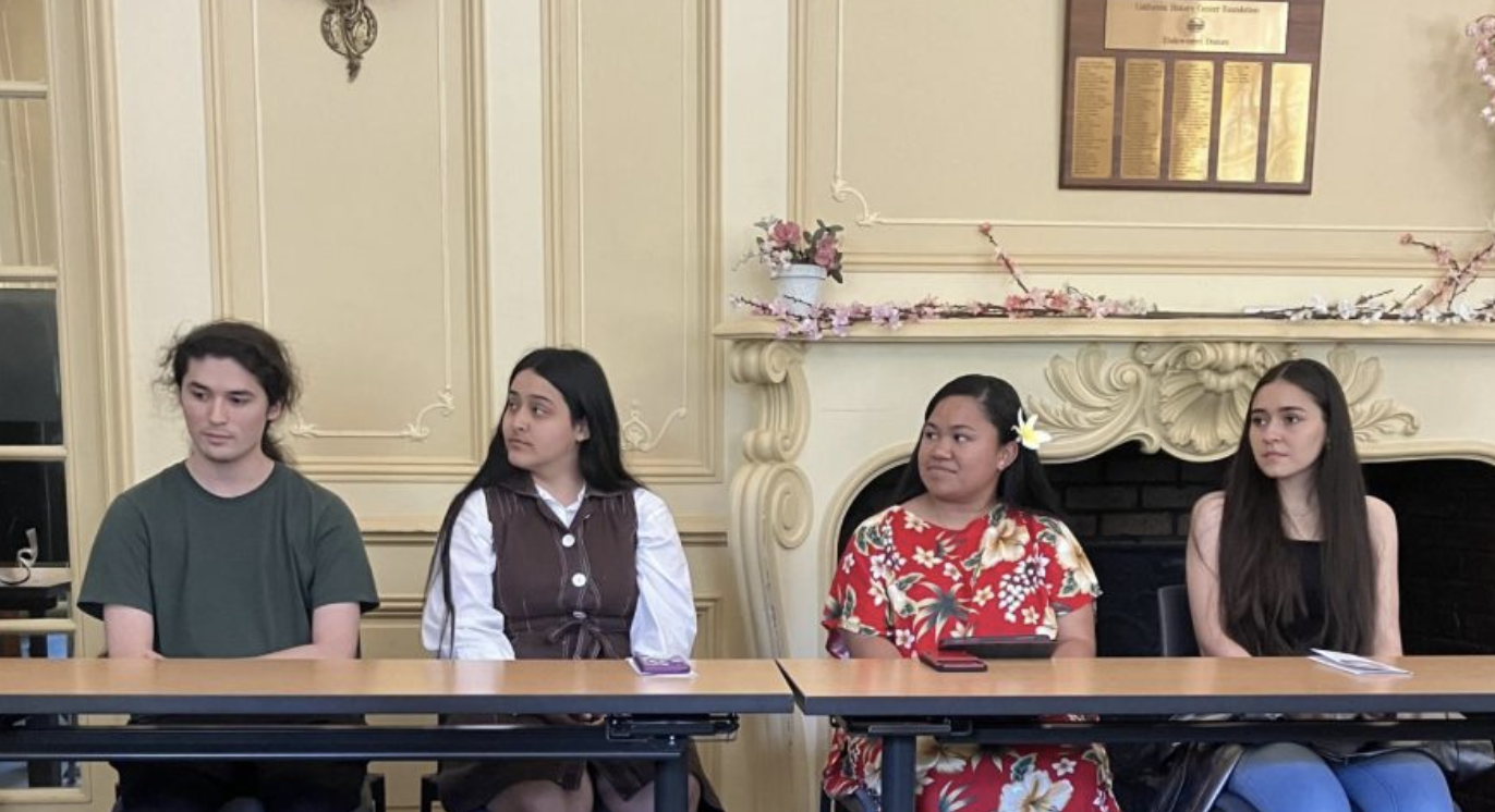 Four people sit at a long table in a decorated room, facing forward.